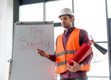 handsome fireman holding red extinguisher while standing near white board with fire safety lettering